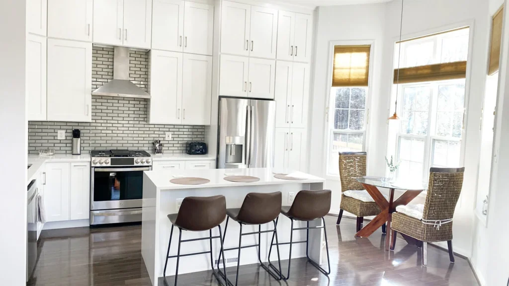 Bright kitchen with white cabinets, island with bar stools, stainless steel appliances, and dining table near windows