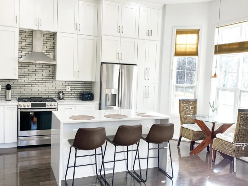 Bright kitchen with white cabinets, island with bar stools, stainless steel appliances, and dining table near windows