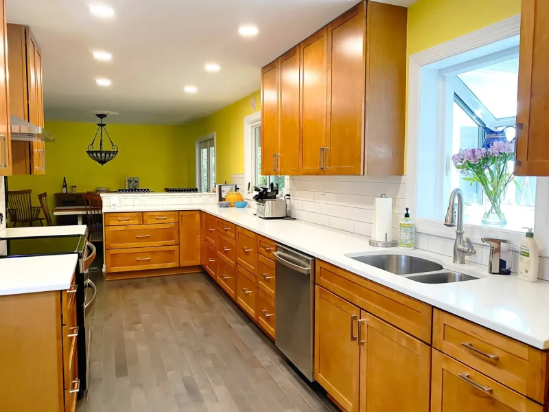 Open concept kitchen with wood cabinets, white countertops, sink under window, and dining area in background